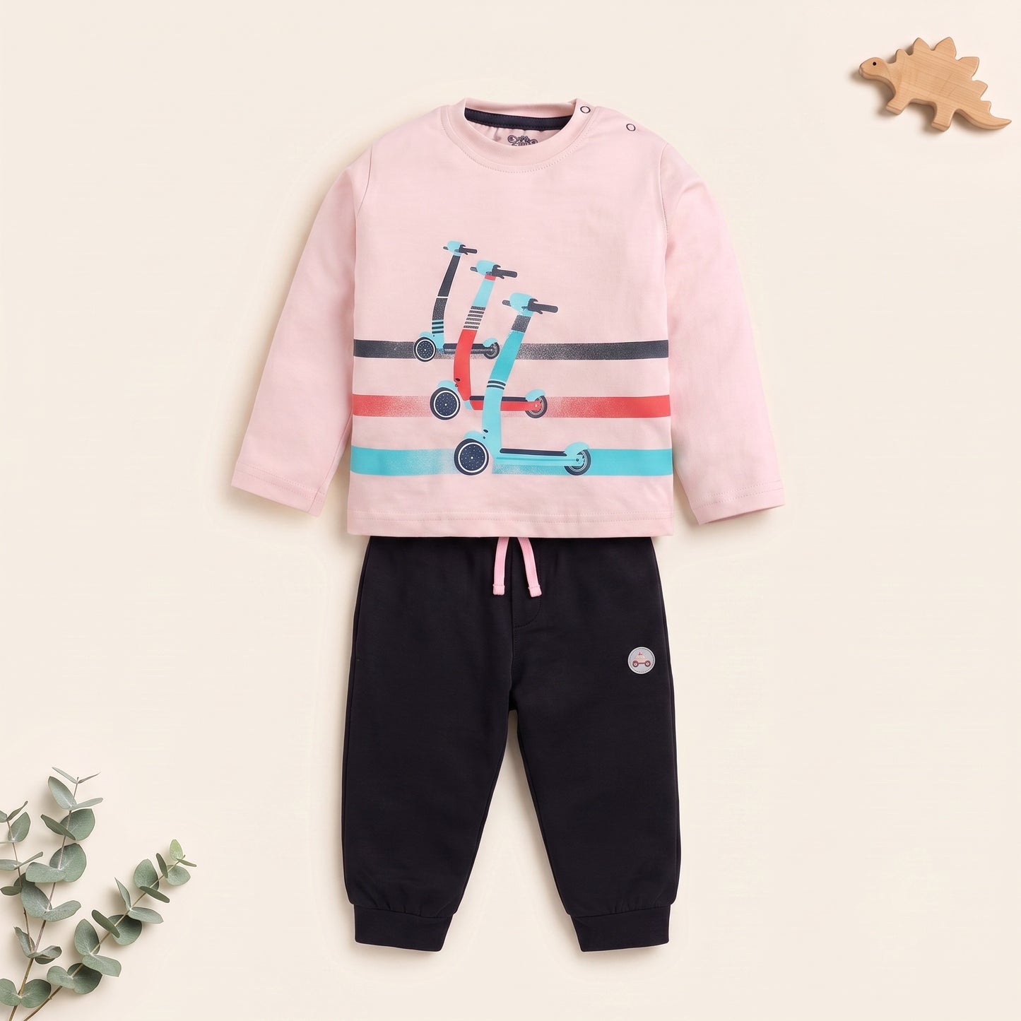 A children's two-piece outfit set featuring a light pink long-sleeve top with a scooter graphic and black jogger pants with a pink drawstring, displayed on a warm cream background with decorative eucalyptus leaves and a wooden dinosaur toy.