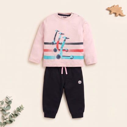 A children's two-piece outfit set featuring a light pink long-sleeve top with a scooter graphic and black jogger pants with a pink drawstring, displayed on a warm cream background with decorative eucalyptus leaves and a wooden dinosaur toy.